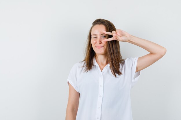 Молодая женщина показывает знак победы на глазах в белой блузке и выглядит довольной