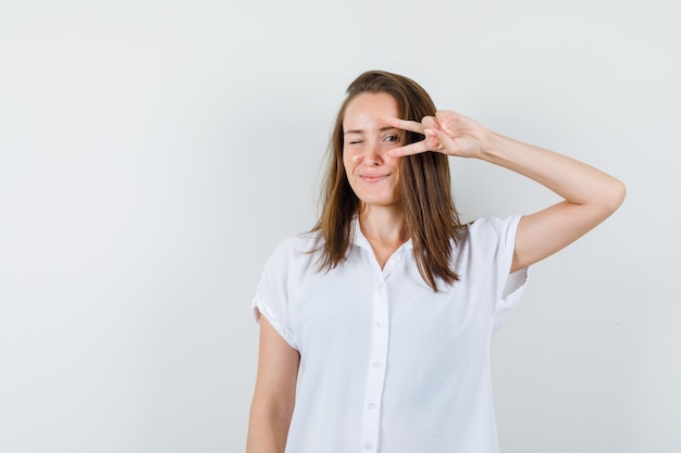 Young female showing victory sign on her eye in white blouse and looking pleased