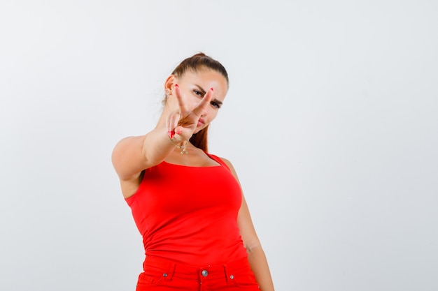 Free photo young female showing victory sign in red tank top, pants and looking lucky , front view.