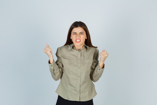 Young female showing winner gesture, clenching teeth in shirt, skirt and looking lucky. front view.