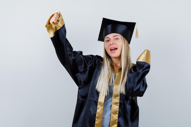 Young female showing winner gesture in graduate uniform and looking lucky.