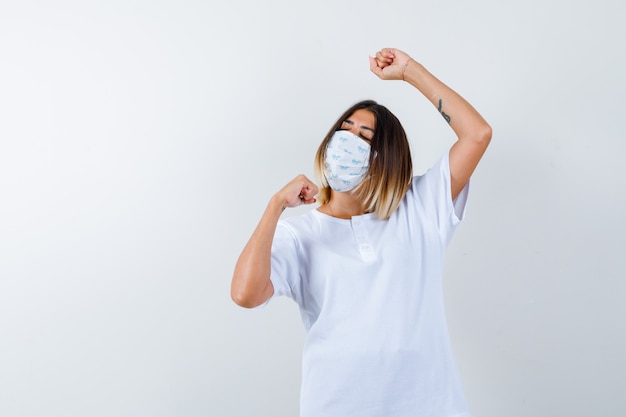 Young female showing winner gesture in t-shirt, mask and looking lucky. front view.