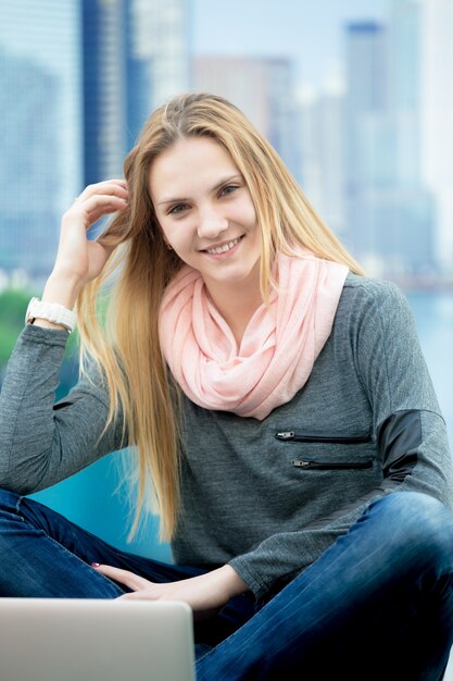 Young female sitting cross-legged working on portable computer, city on the background