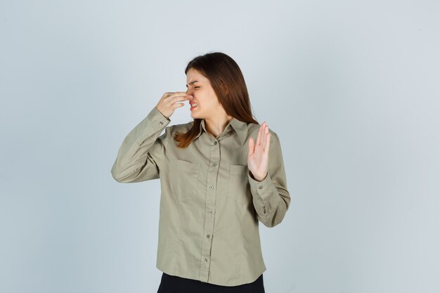 Young female smelling something awful, pinching nose, showing stop sign in shirt, skirt and looking disgusted , front view.