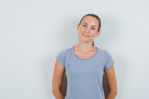 Молодая женщина улыбается с руками на спине в серой футболке, вид спереди.