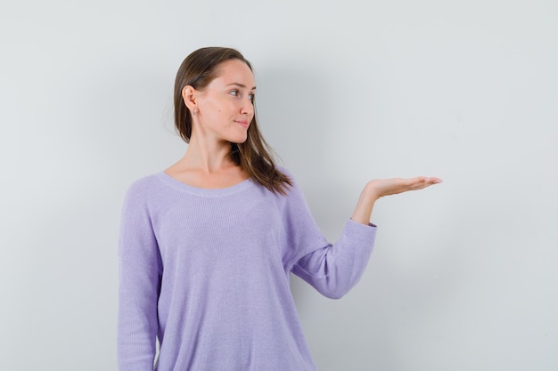 Young female spreading her open palm aside in lilac blouse and looking glad 