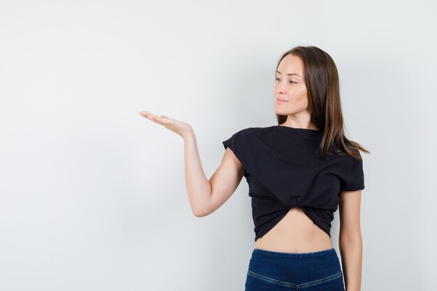 Young female spreading palm aside in black blouse, pants and looking confident