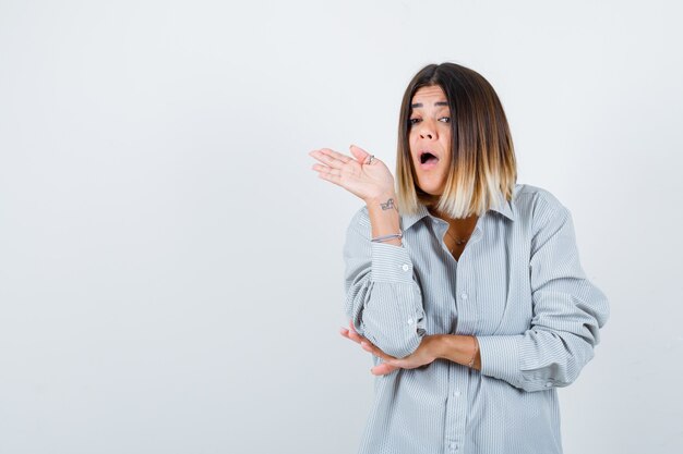 Young female spreads palm out while showing something in oversized shirt and looking perplexed. front view.