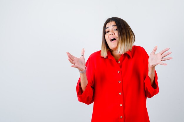 Young female spreads palms out in red oversized shirt and looking amazed , front view.