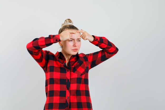 Young female squeezing her pimple on forehead in checked shirt , front view.