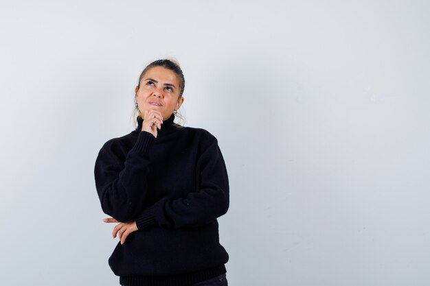 Молодая женщина, стоящая в позе мышления в черном свитере с высоким воротом и задумчивая, вид спереди.