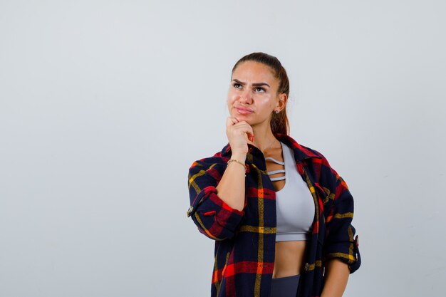 Young female standing in thinking pose in crop top, checkered shirt, pants and looking pensive. front view.