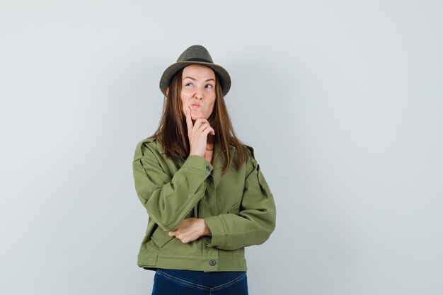 Young female standing in thinking pose in jacket, pants, hat and looking hesitant. front view.