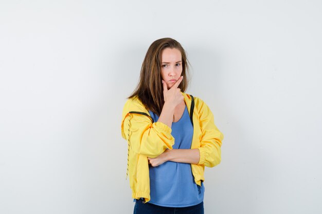 Молодая женщина, стоящая в позе мышления в футболке, куртке и серьезная, вид спереди.