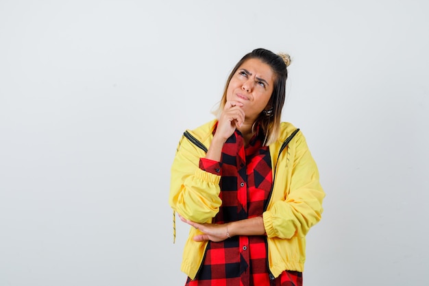 Young female standing in thinking pose while looking up in checkered shirt, jacket  and looking puzzled , front view.