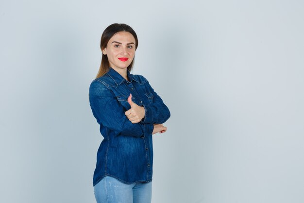 Young female standing with crossed arms while showing thumb up in denim shirt and jeans and looking confident