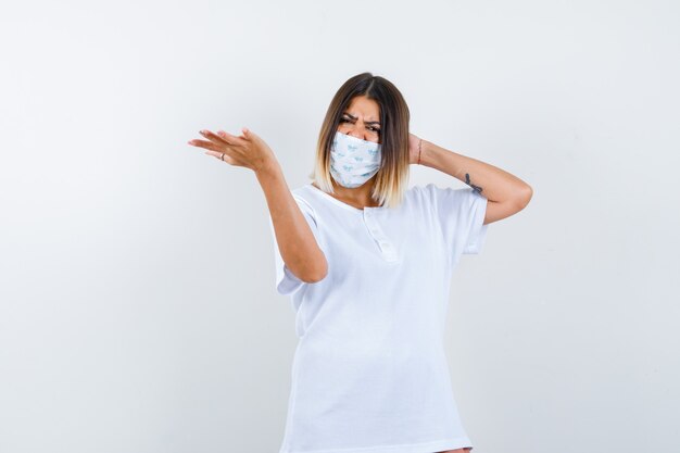 Young female stretching hand in questioning gesture in t-shirt, mask and looking hesitant , front view.