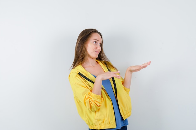Young female stretching hands to show something in t-shirt, jacket and looking elegant, front view.