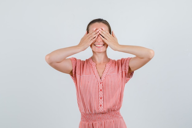 Young female in striped dress covering eyes with hands and looking excited , front view.