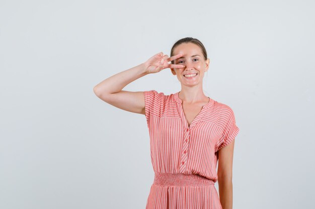 Young female in striped dress showing v-sign near eye and looking happy , front view.