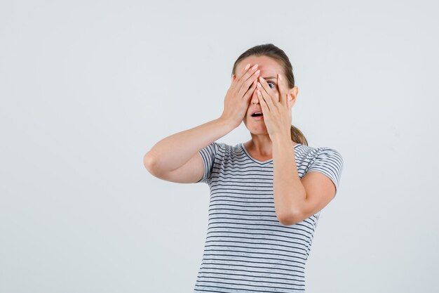 Young female in striped t-shirt looking through fingers and looking scared , front view.