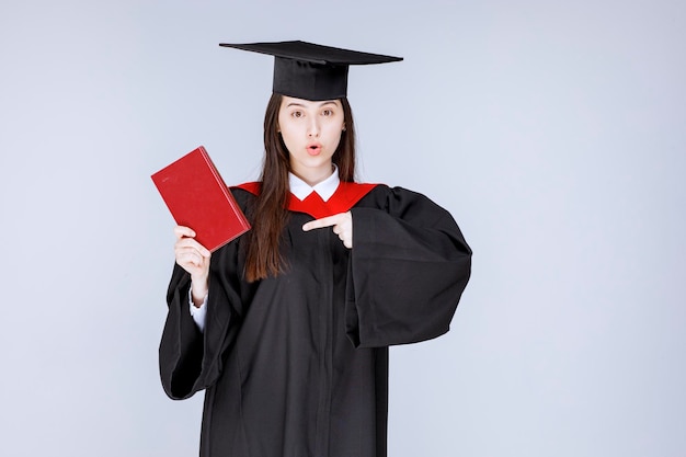 Young female student in academic gown pointing at book. High quality photo