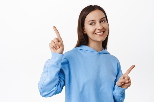 Young female student in blue hoodie, pointing sideways, two choices ways, showing variants of items on sale, smiling and lookin left at one side, standing over white background.