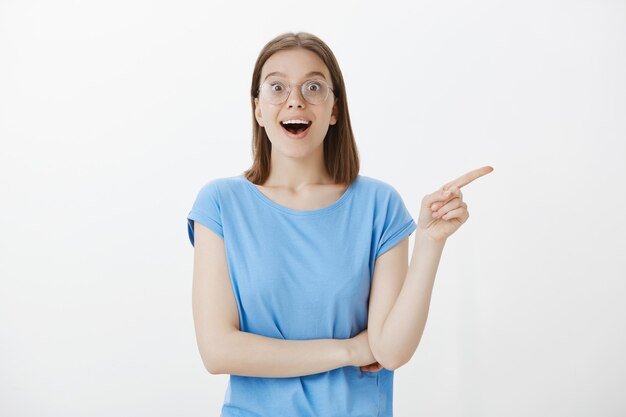 Young female student in glasses smiling and pointing finger right, looking intrigued