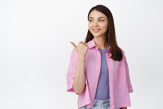 Young female student pointing and looking left with hopeful smiling face expression standing over white background