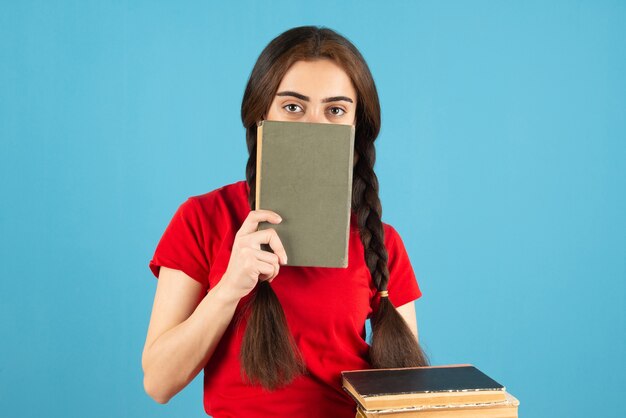 Young female student in red t-shirt hiding behind book on blue wall.