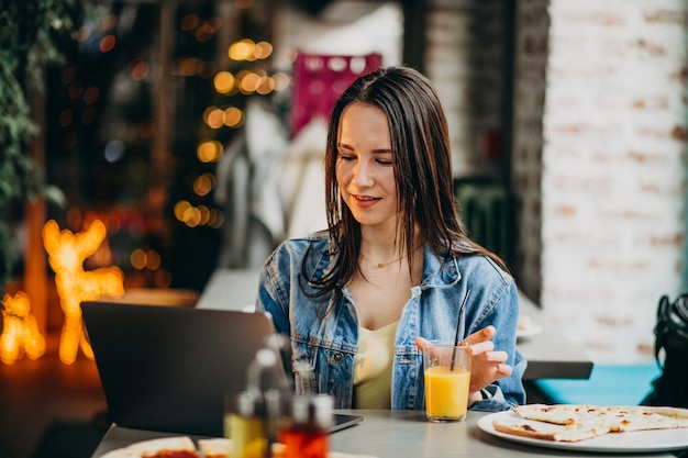 Free photo young female student working on laptop in bar and eating pizza