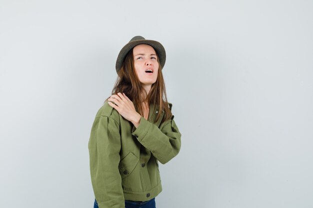 Young female suffering from shoulder pain in jacket, pants, hat and looking fatigued. front view.