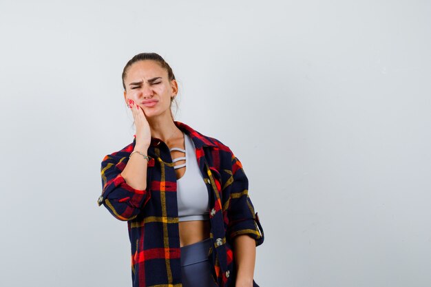 Young female suffering from toothache in crop top, checkered shirt, pants and looking unwell , front view.