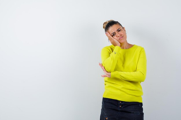 Young female in sweater, denim skirt leaning cheek on hand and looking thoughtful 