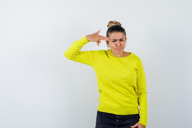 Young female in sweater, denim skirt shooting herself with hand gun and looking bored 