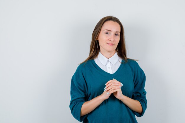 Молодая женщина в свитере над рубашкой держит переплетенные пальцы на груди и выглядит разумно, вид спереди.