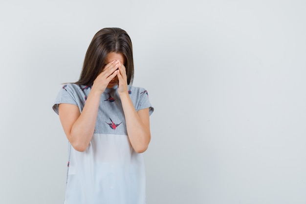 Young female in t-shirt bending head down with hands on face and looking sad , front view.