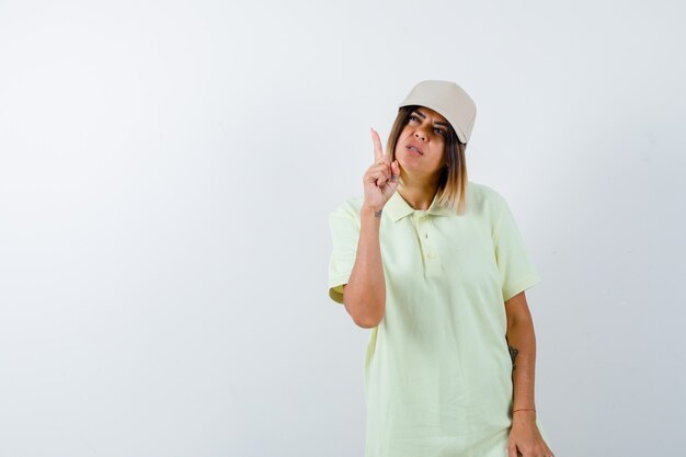 Young female in t-shirt, cap pointing up and looking hesitant , front view.