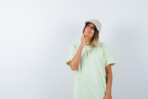T- 셔츠에 젊은 여성, 생각 포즈에 서있는 모자와 사려 깊은, 전면보기를 찾고.