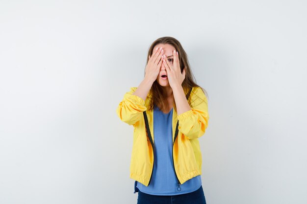 Young female in t-shirt, jacket looking through fingers and looking scared , front view.