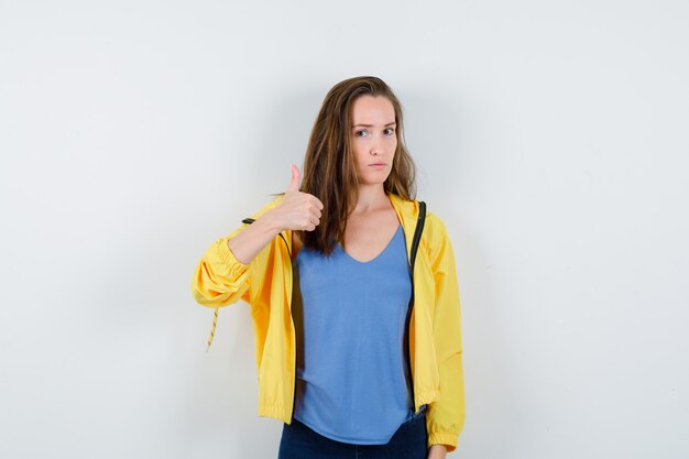 Young female in t-shirt, jacket showing thumb up and looking confident, front view.