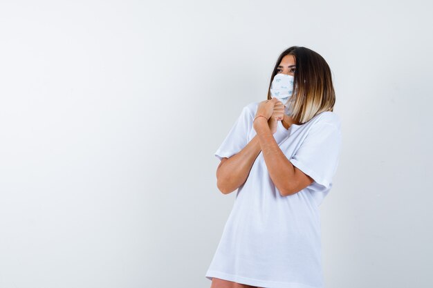 Young female in t-shirt, mask holding clenched hands while looking up and looking amazed , front view.