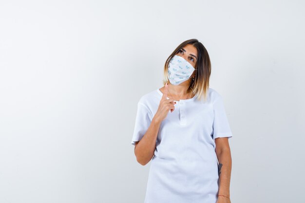 Young female in t-shirt, mask pointing up and looking thoughtful , front view.