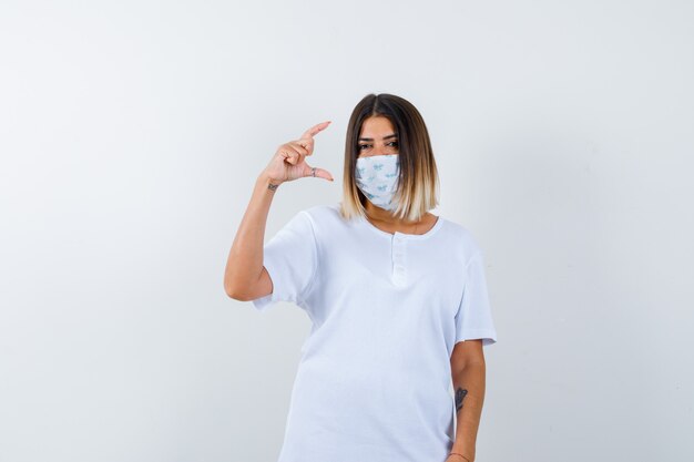 Молодая женщина в футболке, маска показывает знак размера и выглядит уверенно, вид спереди.