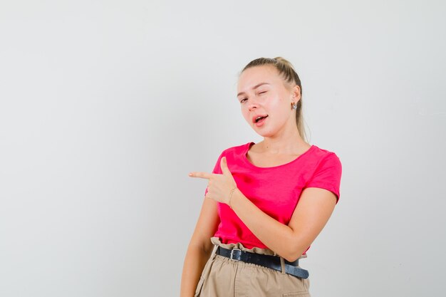 Young female in t-shirt, pants pointing aside, winking eye and looking confident , front view.