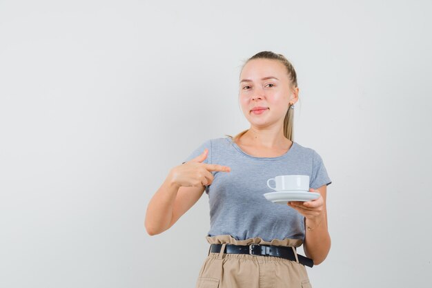 Молодая женщина в футболке, штаны, указывая на чашку напитка и рад, вид спереди.