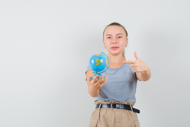 Young female in t-shirt, pants pointing at globe model and smiling , front view.