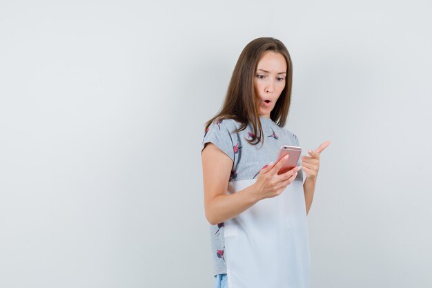 Young female in t-shirt pointing at mobile phone and looking amazed , front view.