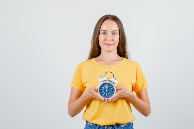 Young female in t-shirt, shorts holding alarm clock and smiling