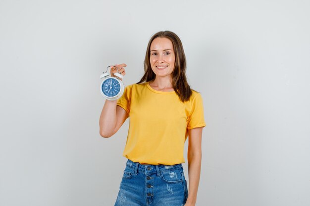 Young female in t-shirt, shorts holding alarm clock and smiling